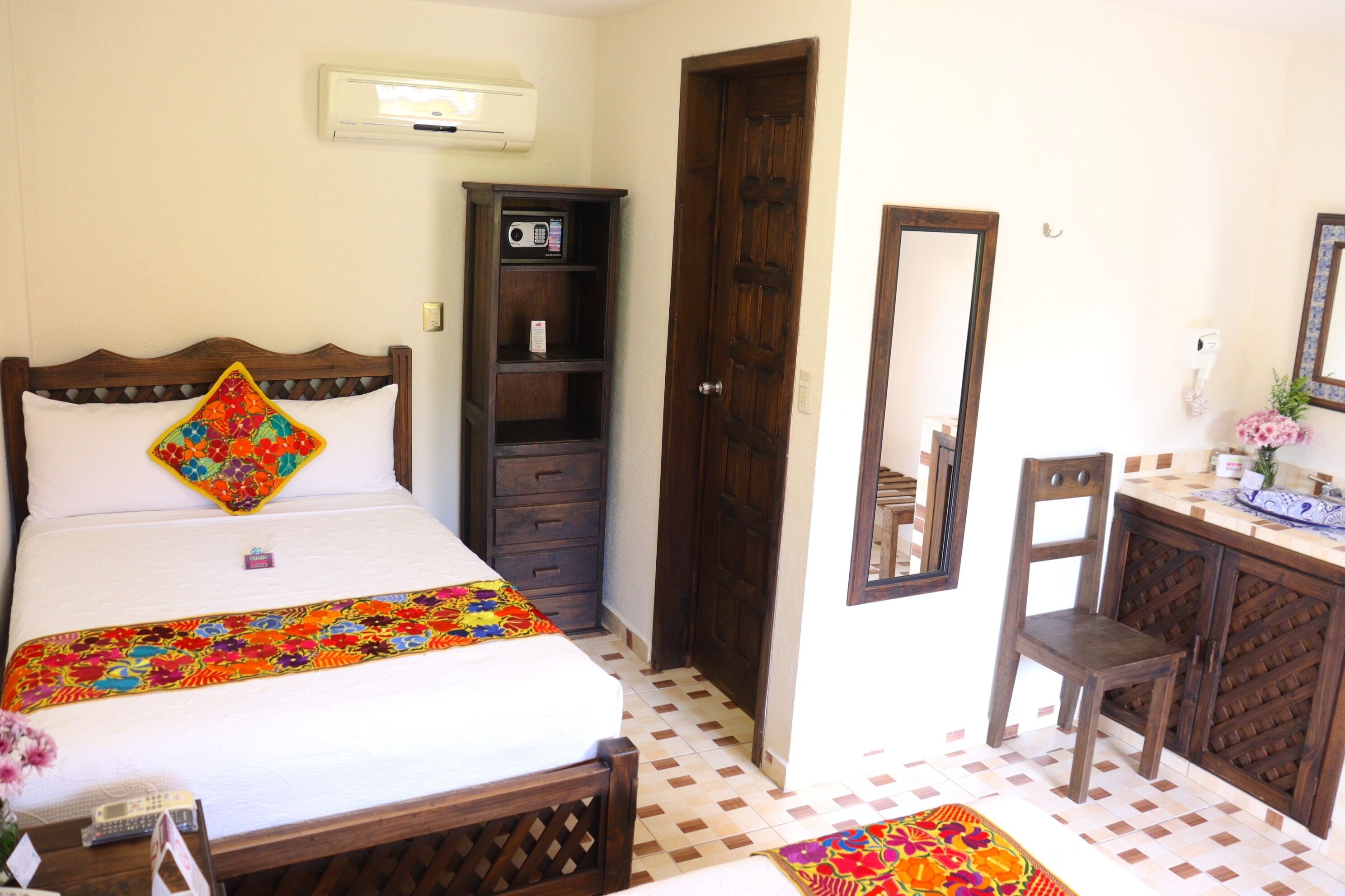 A hotel room with a bed, a wooden wardrobe, a desk with a chair, and a mirror on the wall.
