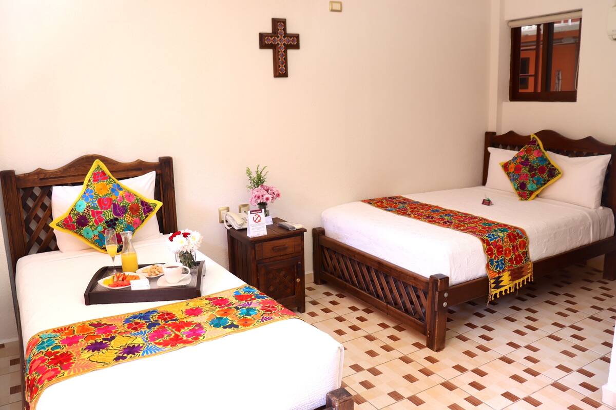 Habitación de hotel con dos camas, cabecero de madera, una cruz en la pared y suelo de baldosas.