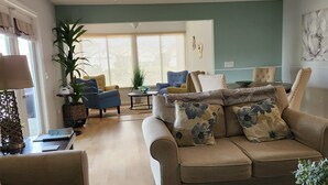 Un salon avec un canapé beige, une table basse et une table à manger. Des chaises sont disposées autour de la table à manger et une plante se trouve sur le rebord de la fenêtre.