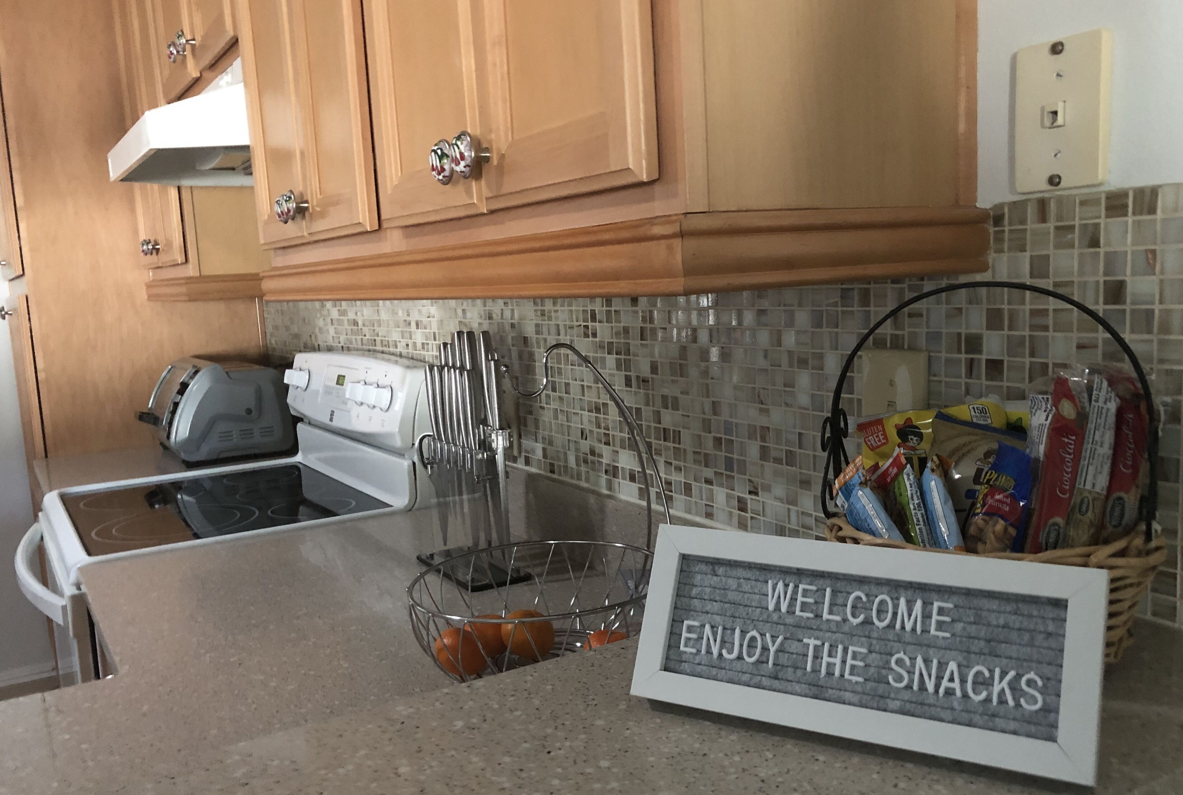 Une cuisine avec des meubles en bois, une hotte de cuisinière en acier inoxydable, un grille-pain, une bouilloire et un panier rempli d’en-cas, accompagné d’une pancarte indiquant : « BIENVENUE, SERVEZ-VOUS ».