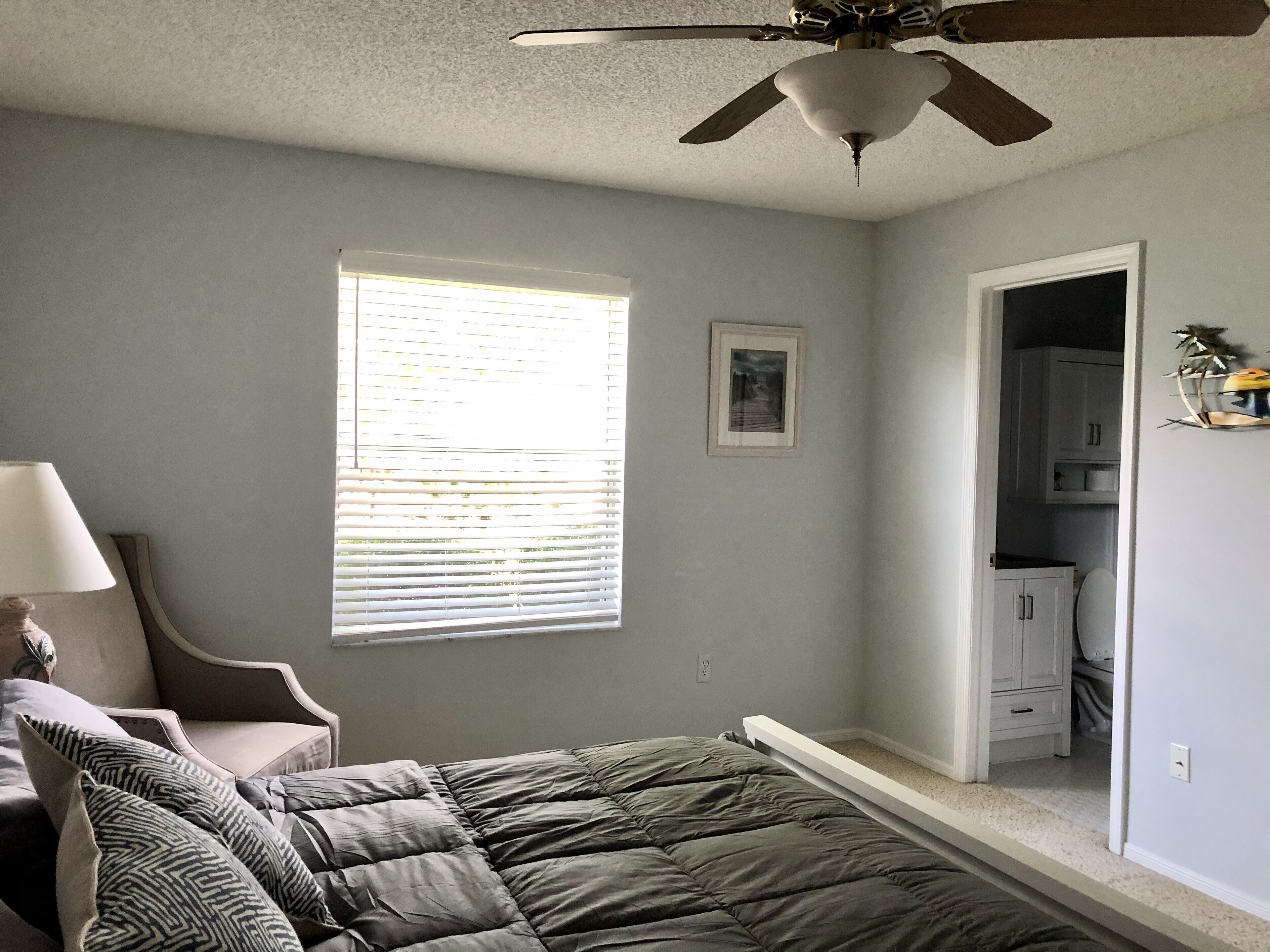 A bedroom with a bed, a ceiling fan, a window with blinds, a chair, and a door leading to a bathroom.