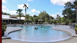 Une piscine entourée de chaises longues et de parasols, bordée de palmiers et dotée d’un espace couvert avec un toit.