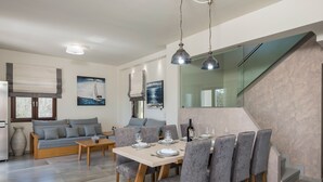 Een moderne woonkamer met een bank, eettafel en stoelen. Er is een trap met een glazen balustrade en een schilderij van een zeilboot aan de muur.