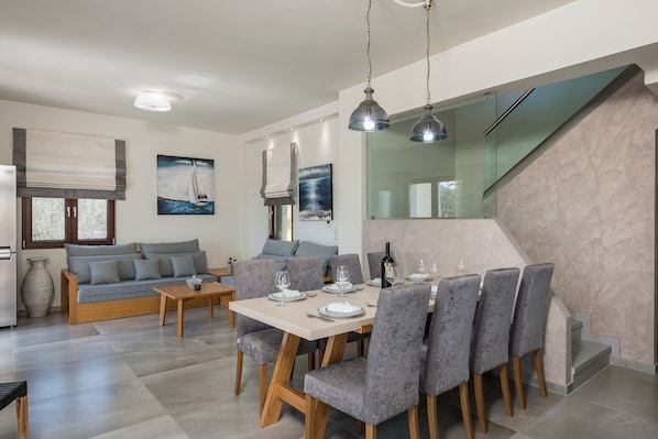 A modern living room with a sofa, dining table set for six, and a staircase with a glass railing.