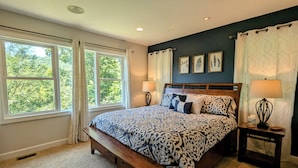 Une chambre avec un grand lit, deux tables de chevet avec des lampes, un banc au pied du lit et une fenêtre avec des rideaux.