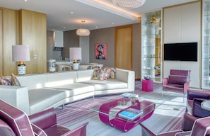 A modern living room with a white sofa, a glass coffee table, and a pink armchair. There is a flat-screen TV mounted on the wall and a decorative shelf with glassware.