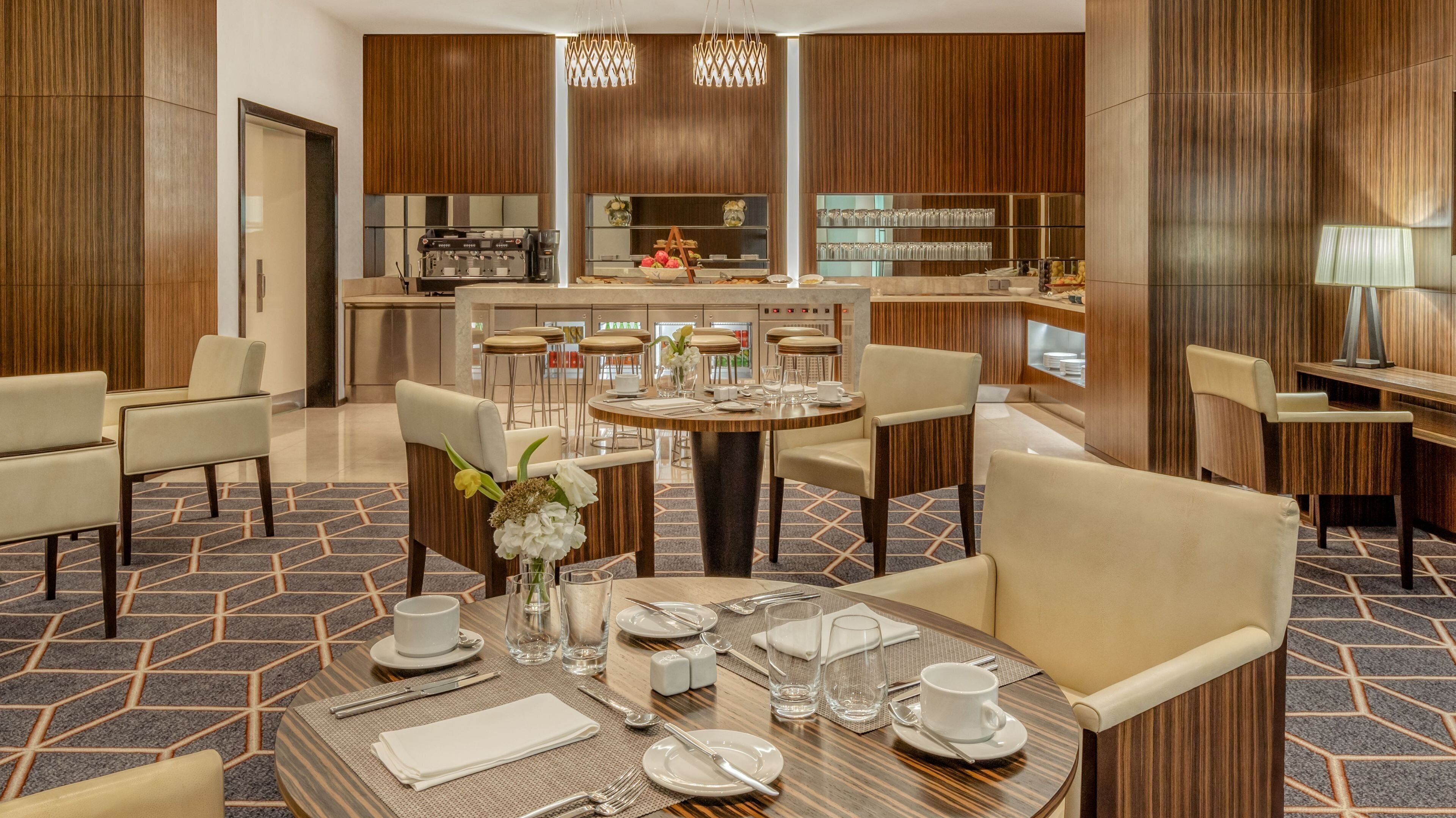 A hotel dining area with a round table set for four, bar stools, and a kitchen in the background.