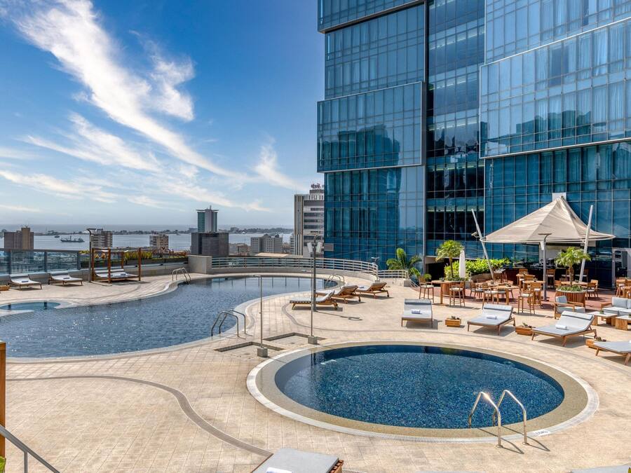 Un área de piscina en la azotea con sillones de descanso y una piscina grande en el centro, rodeada de edificios modernos.