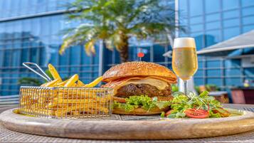 Comida que incluye una hamburguesa, papas fritas y una ensalada, con una copa de champán, sobre una mesa al aire libre, con un edificio moderno al fondo.