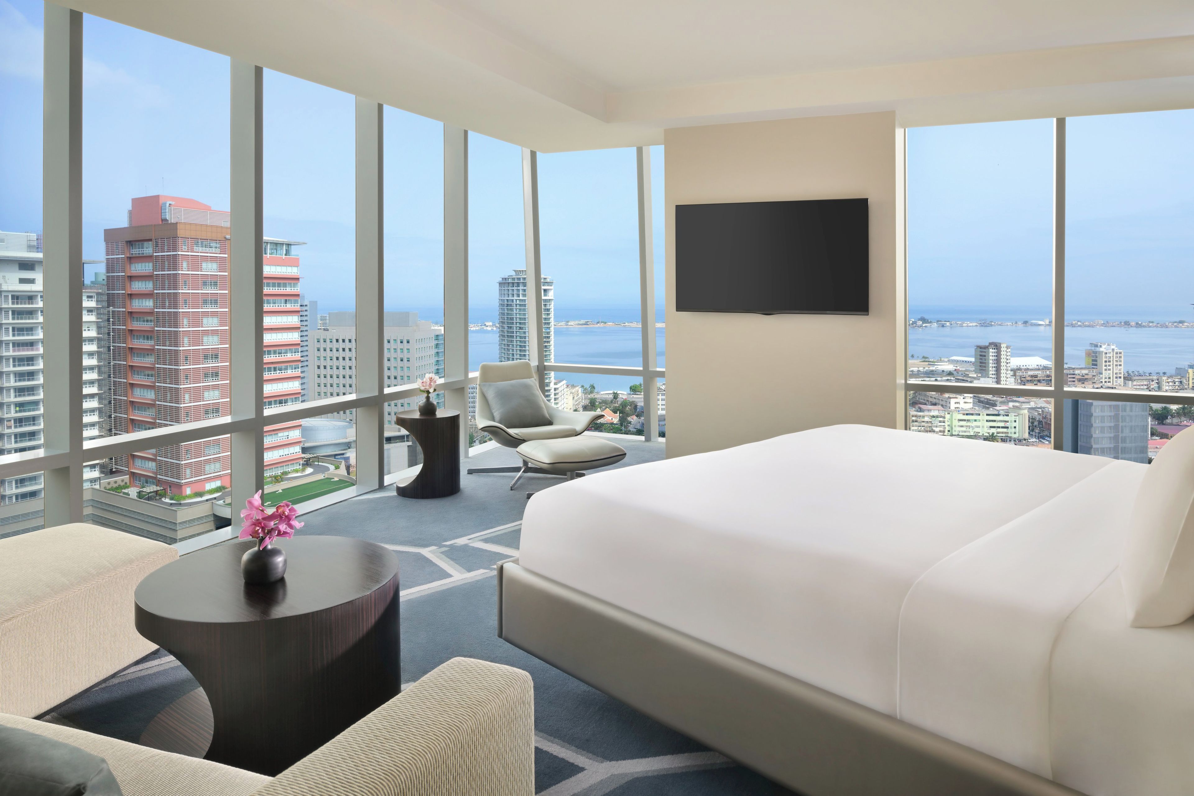 A hotel room with a large bed, a chair, a small table with a vase, and a TV on the wall.