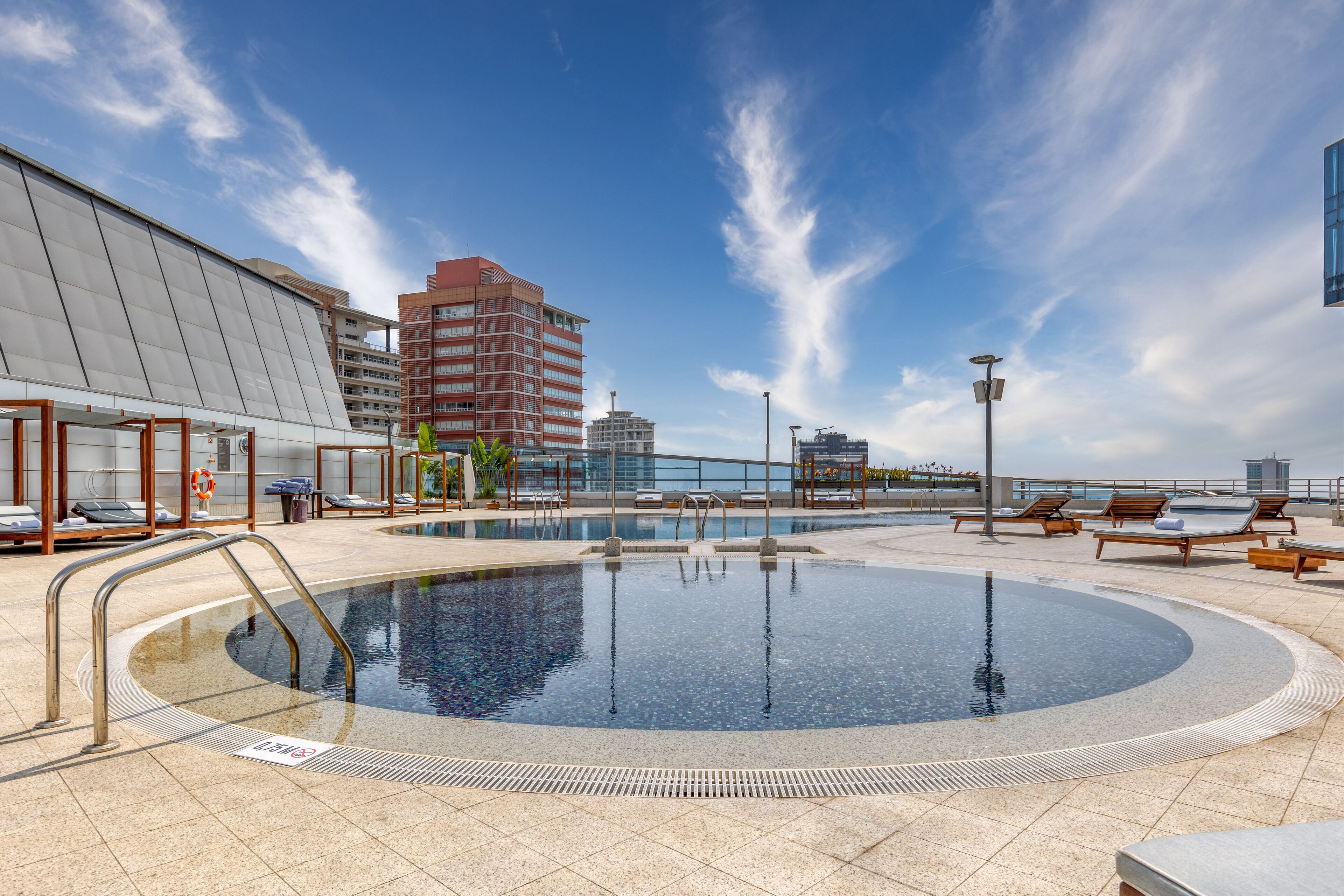A rooftop swimming pool with a clear view of the city skyline, lounge chairs, and a modern building with a glass facade.
