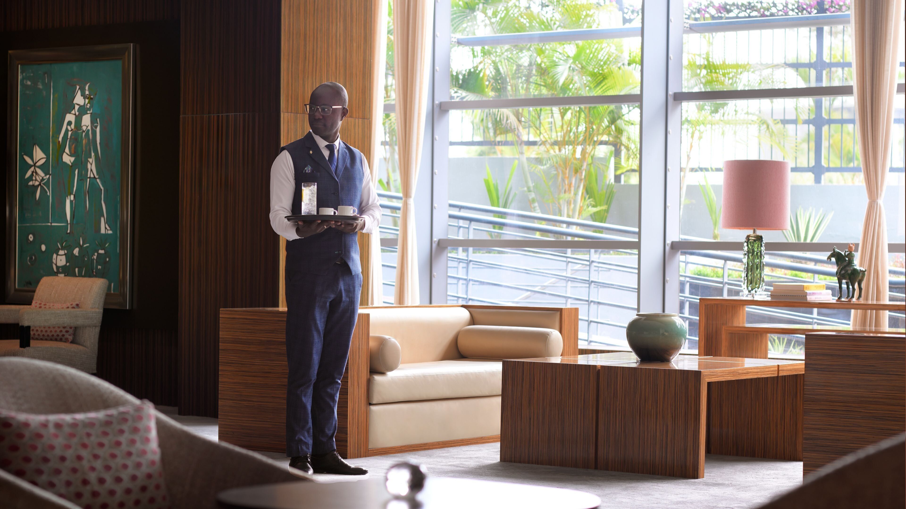 A man in a vest holding a tray with cups and a glass, standing in a room with a large window, a sofa, and a table with a vase and figurine.