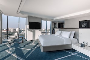 A modern hotel room with a large bed, a flat-screen TV, a seating area with a chair, and a view of the city through large windows.
