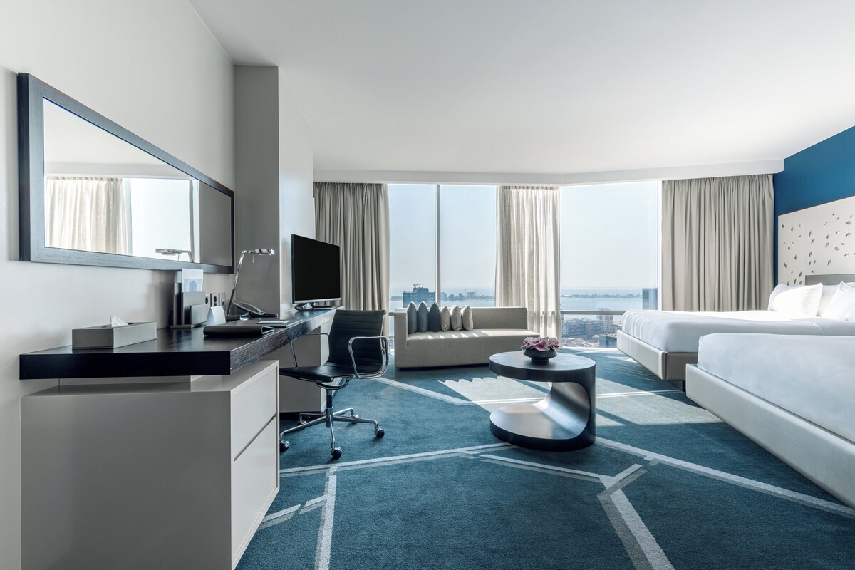 Een moderne hotelkamer met een groot bed, een slaapbank, een bureau met een computer en een groot raam met uitzicht op de stad.