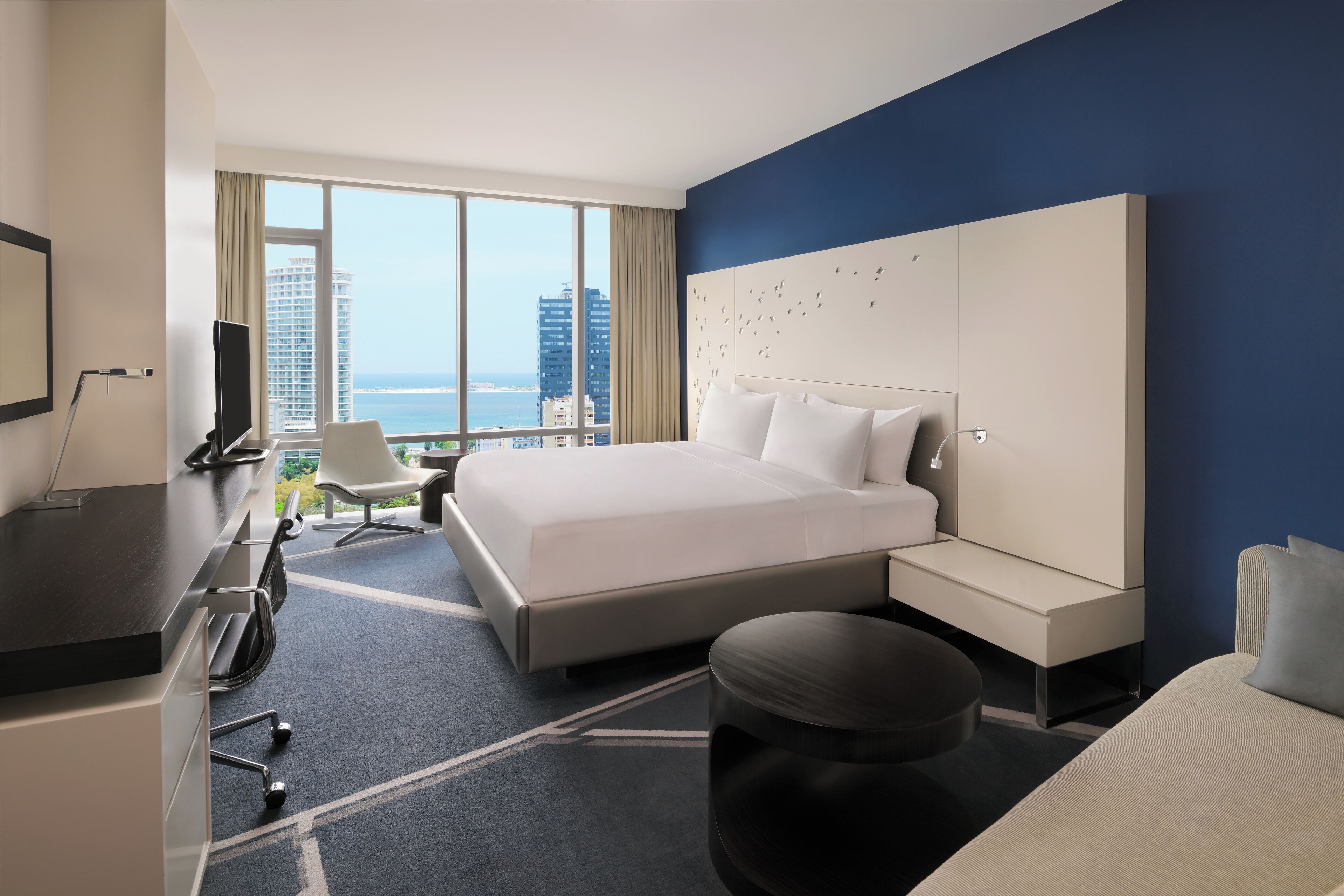 A modern hotel room with a large bed, a desk, a chair, and a view of the cityscape.