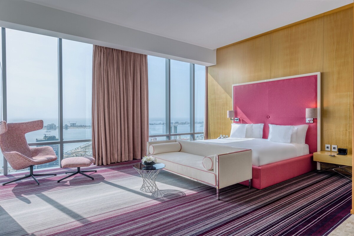 Een moderne hotelkamer met een groot bed, een roze hoofdeinde, een sofa, een stoel en uitzicht op de stad door grote ramen.