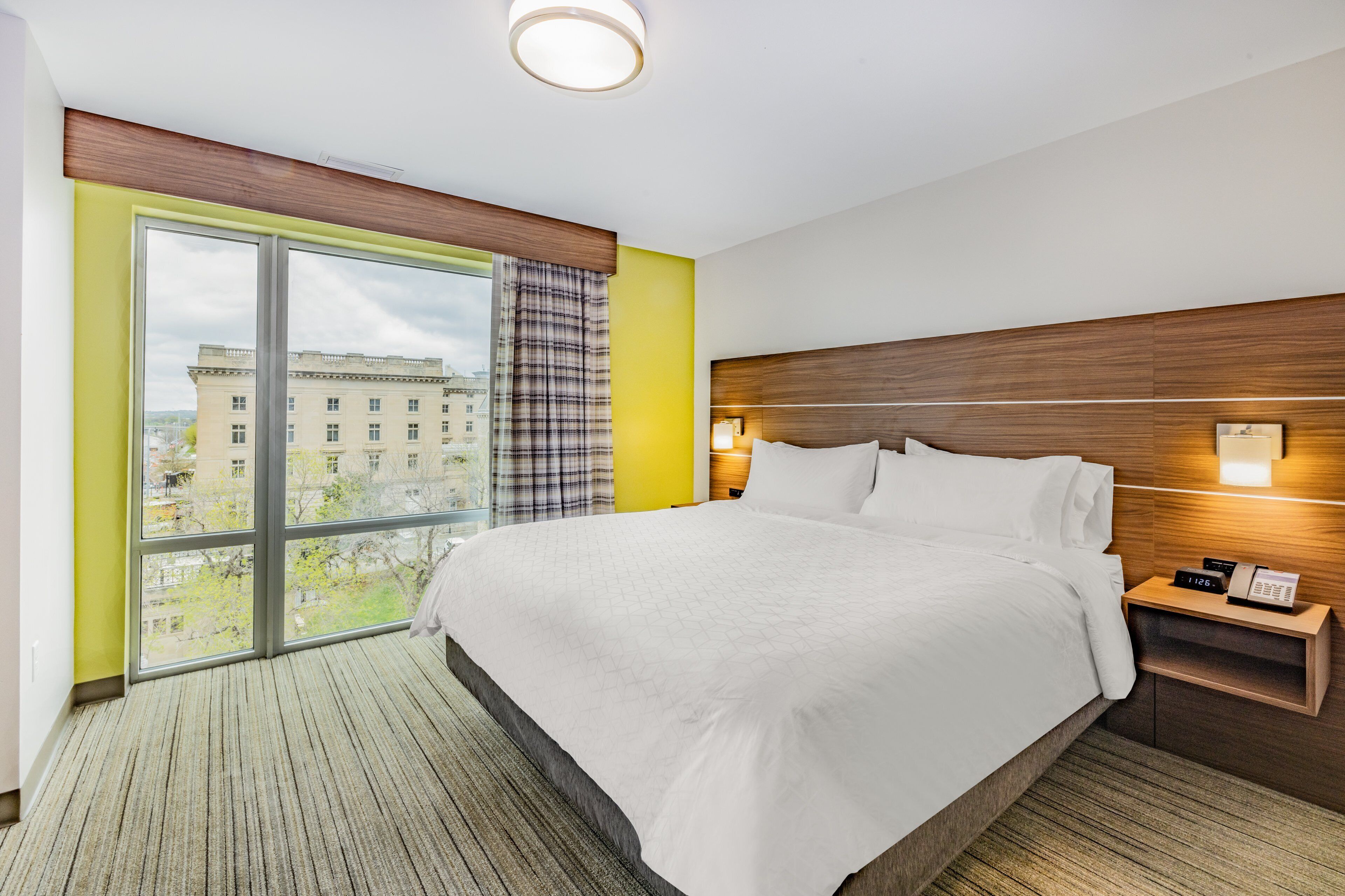 Une chambre d’hôtel avec un grand lit, une table de chevet avec un téléphone, une fenêtre donnant sur des immeubles et un luminaire au plafond.