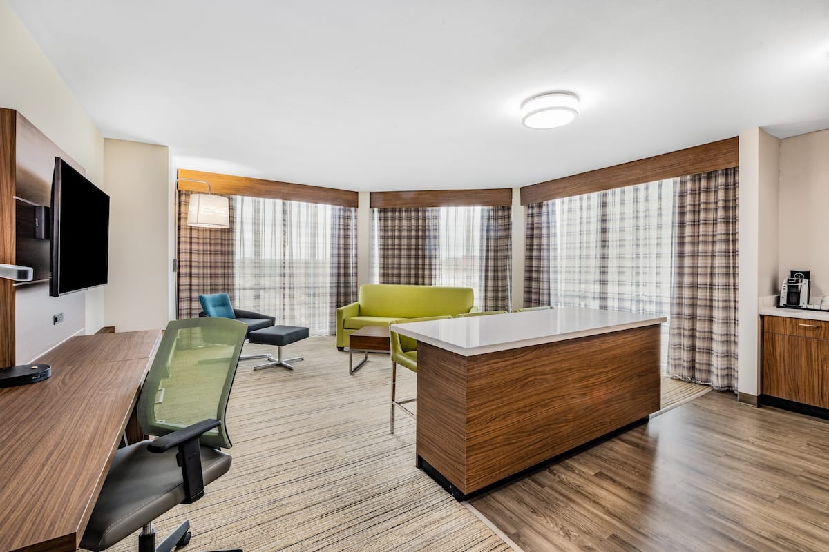 A modern hotel room with a wooden desk, a green chair, a sofa, and a large window with curtains.
