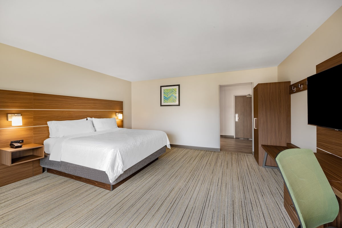 A hotel room with a large bed, a desk, a chair, a TV, and a door leading to another room.