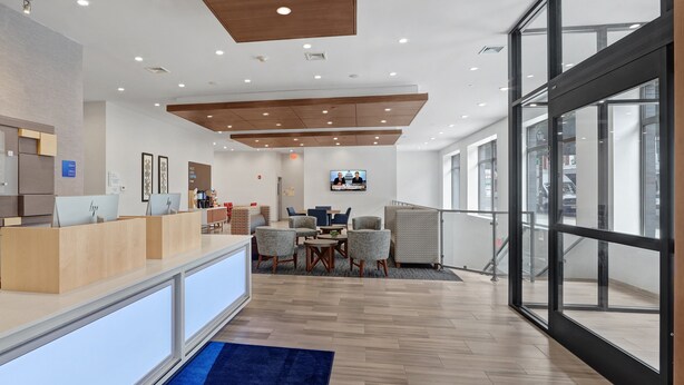 A modern reception area with a wooden reception desk, seating area with chairs, a television screen, and large windows providing natural light. - Hotel 1200 (Brooklyn)