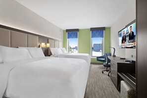 Ein Hotelzimmer mit zwei Betten, einem Schreibtisch, einem Sessel, einem Fernseher und Blick auf die Stadt.