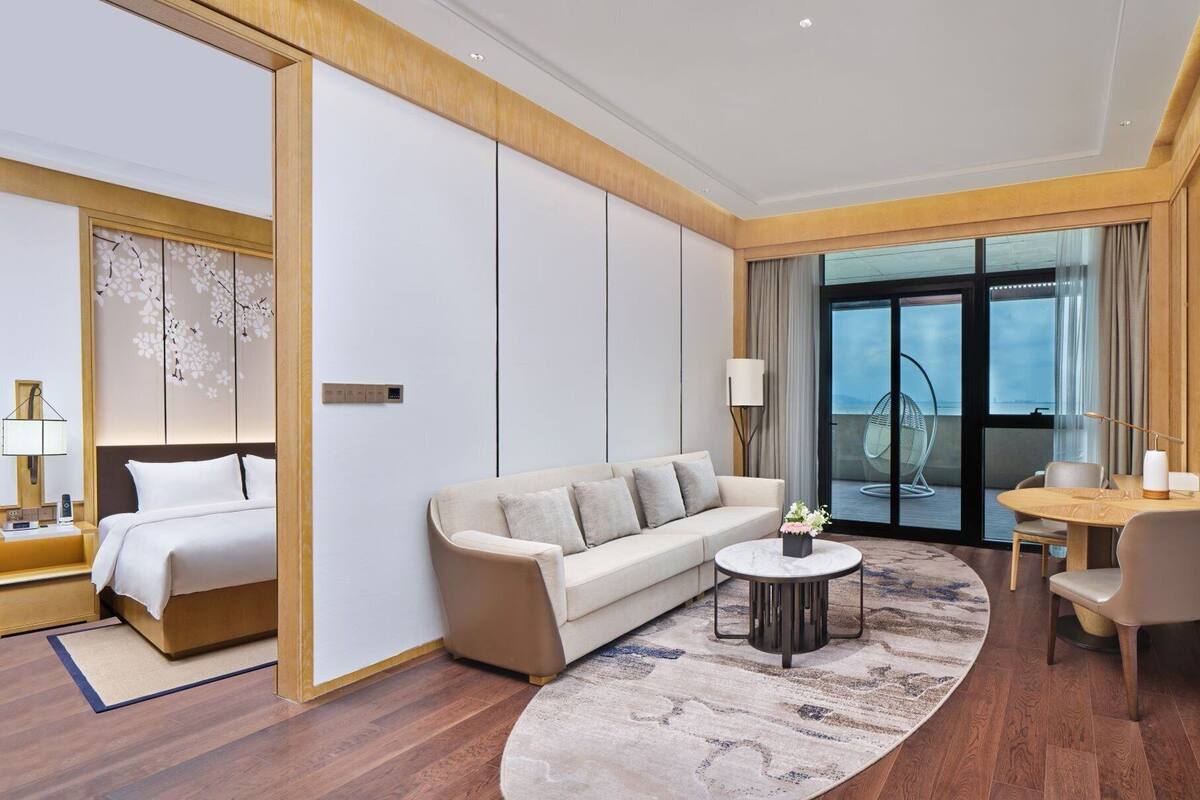 A modern hotel room with a bed, sofa, coffee table, and a dining area with a TV.