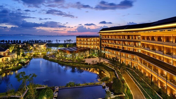 A hotel with a large pool, multiple floors, and a view of the water body in the background. - HUALUXE Suzhou Bay Hot Spring Resort by IHG (Suzhou)