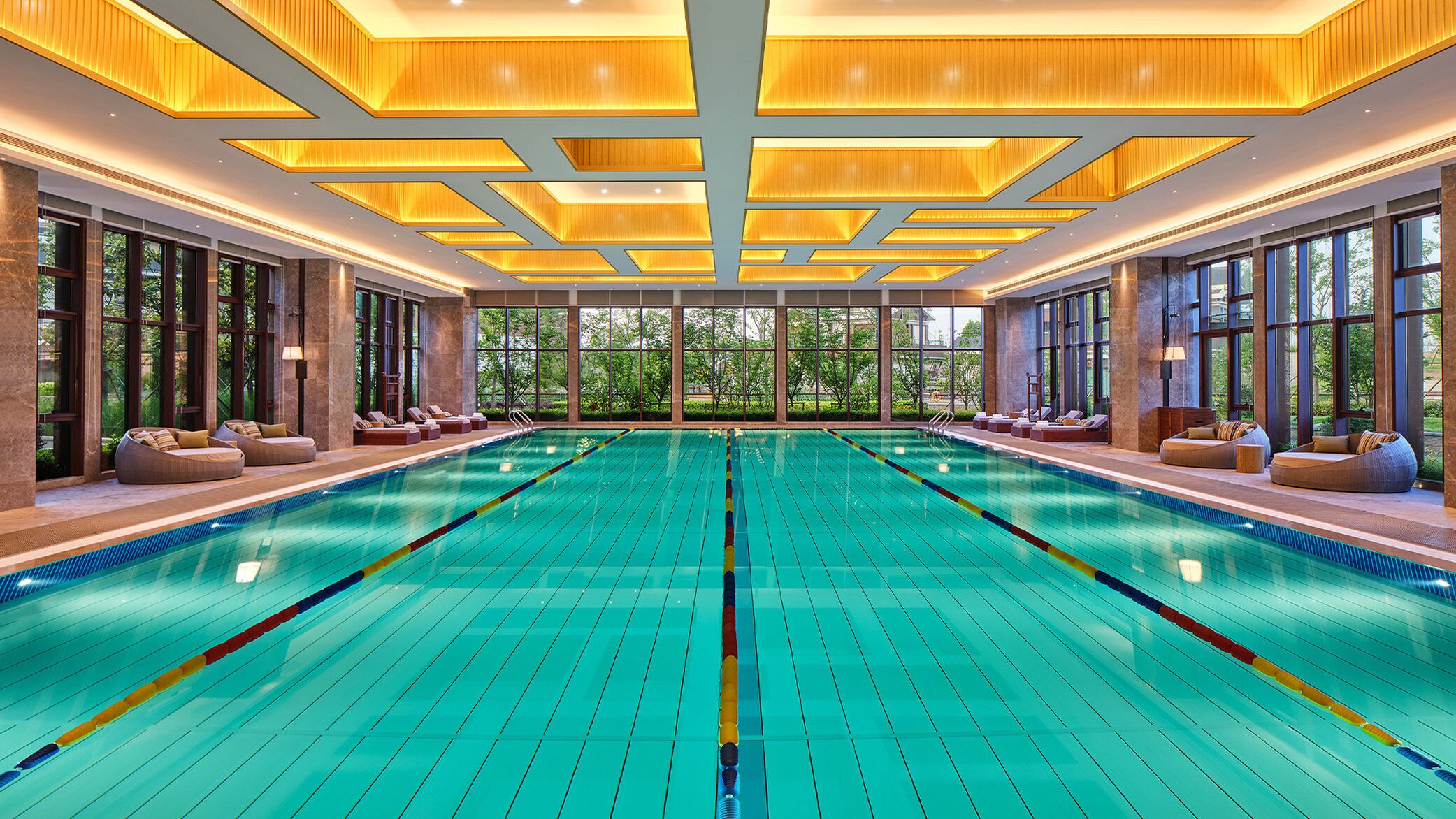 A large indoor swimming pool with clear lane dividers, large windows, and seating areas along the sides.