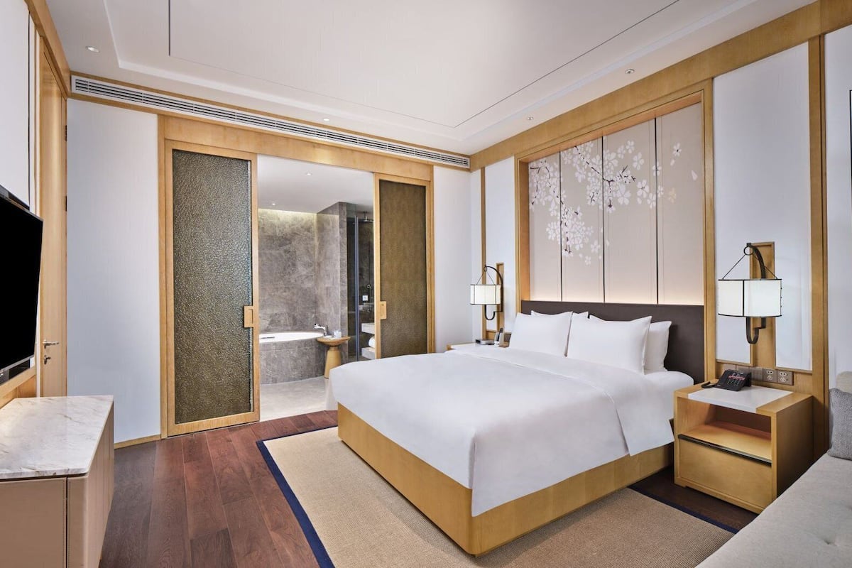 A modern hotel room with a large bed, a TV, a bathroom with a shower, and a desk with a lamp.