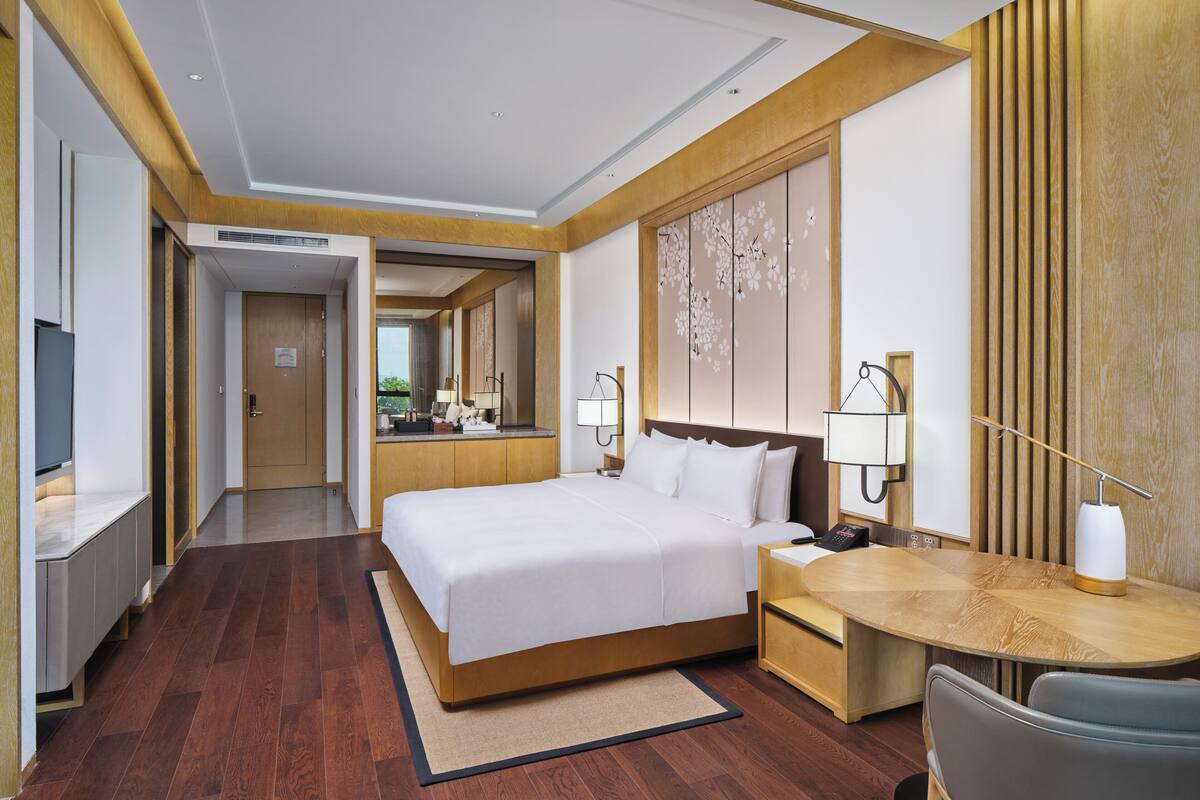 A hotel room with a large bed, a desk with a chair, a TV, and a bathroom visible in the background.