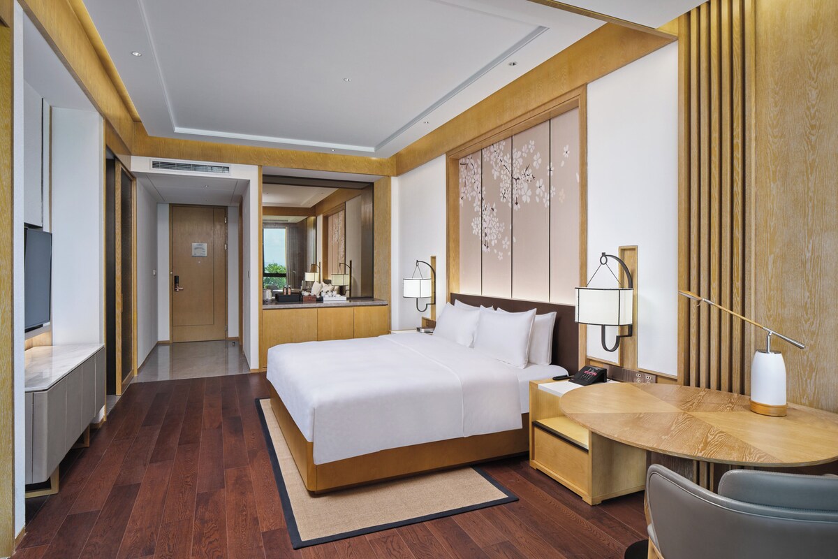 A hotel room with a large bed, a desk with a chair, a TV, and a bathroom visible in the background.