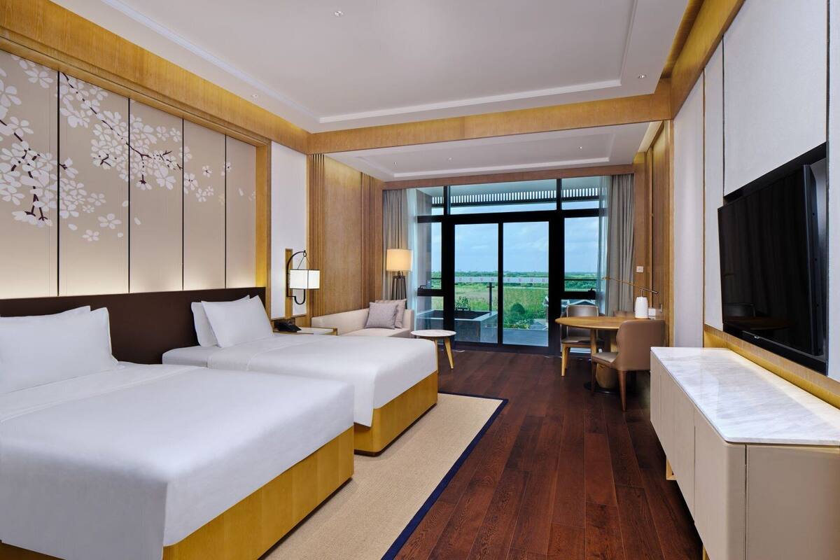 A hotel room with two beds, a desk, a TV, and a large window offering a view of the outdoors.