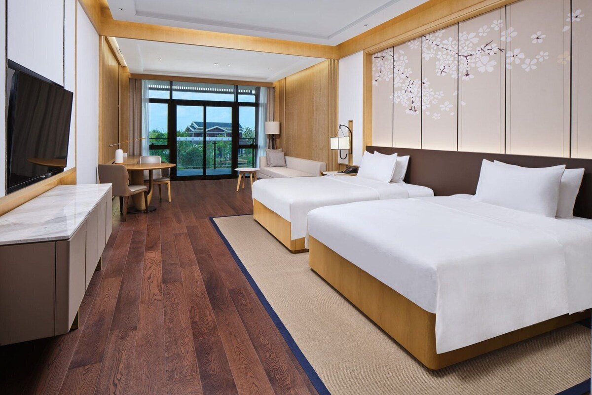 A hotel room with two beds, a desk, a TV, and a large window with a view of greenery.