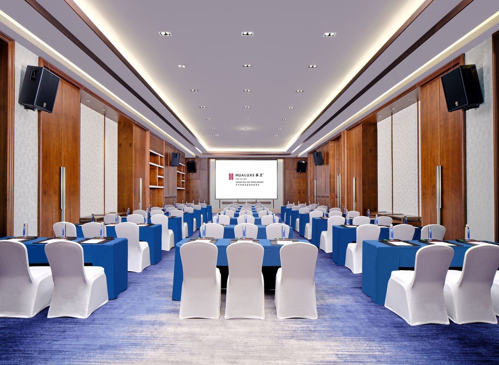 Une salle de conférence avec des rangées de tables et de chaises, équipée de microphones et de projecteurs, et un grand écran à l’avant.