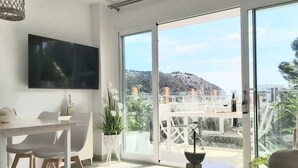 Un salon moderne doté d’un téléviseur à écran plat, d’un coin repas avec des chaises, une table ronde et un balcon offrant une vue sur une colline.