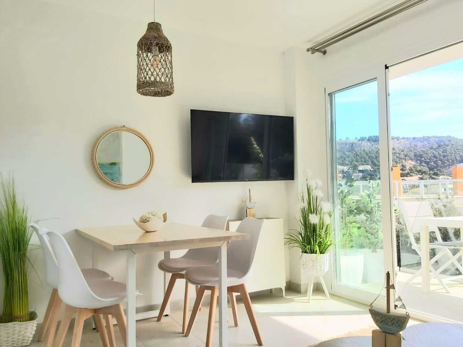 Un espace de repas moderne, avec une table et des chaises, un luminaire suspendu, un téléviseur fixé au mur et une grande fenêtre donnant sur un paysage vallonné.