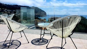 Un balcon avec deux fauteuils en osier blanc et une petite table, d’où l’on peut admirer une ville côtière et la mer.