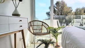 Une chambre avec une grande fenêtre, un lit, un bureau avec des tiroirs, une chaise en osier et deux plantes en pot.