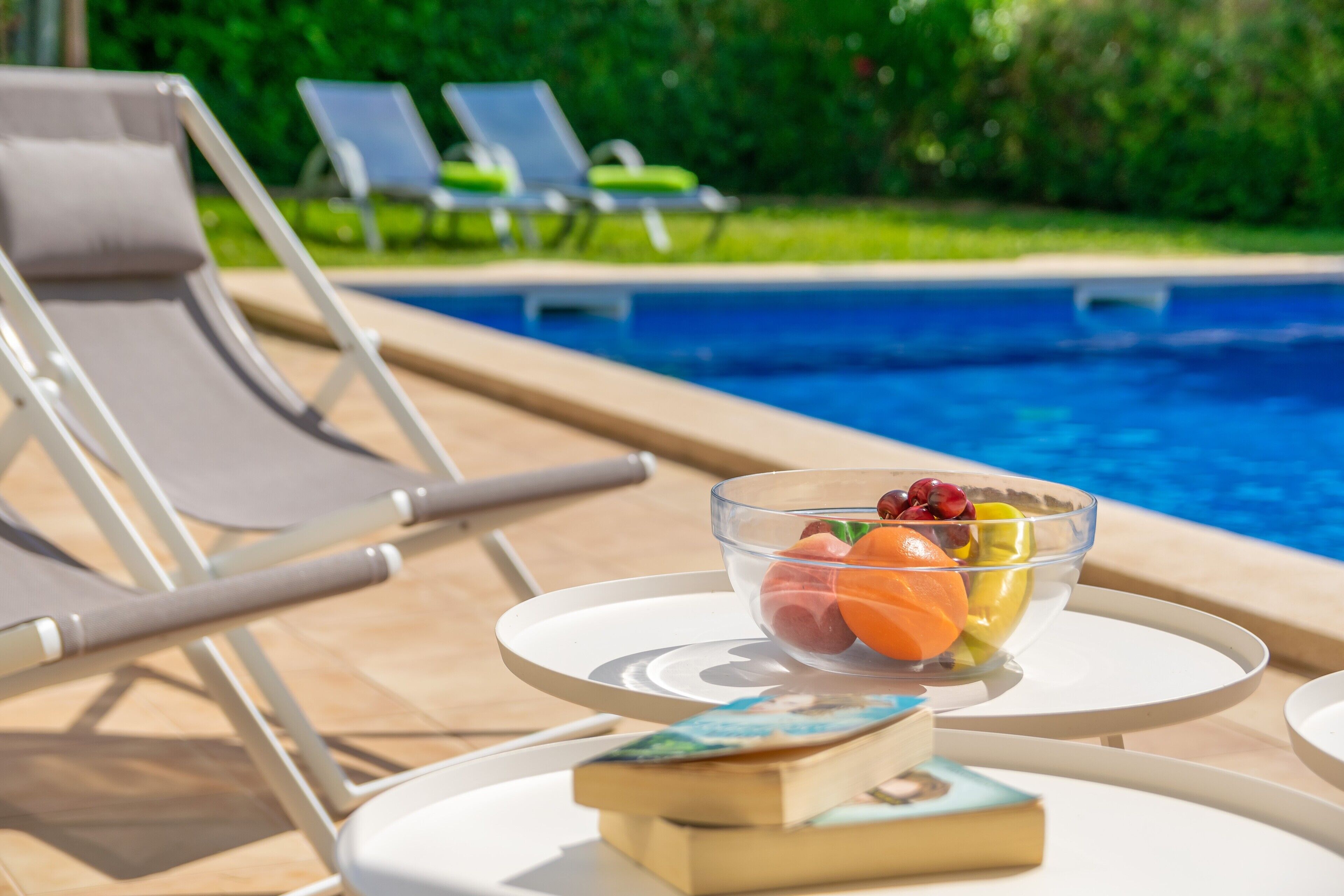 Un espace aménagé au bord de la piscine, avec des chaises longues et une table sur laquelle se trouvent un saladier rempli de fruits et un livre.