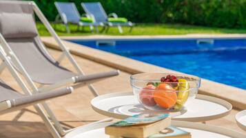 Un espace aménagé au bord de la piscine, avec des chaises longues et une table sur laquelle se trouvent un saladier rempli de fruits et un livre.