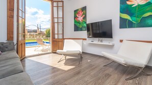 Un salón amplio con un gran ventanal corredera que da a una zona de piscina, un televisor de pantalla plana montado en la pared y dos sillas blancas.