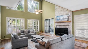 A spacious living room with a fireplace, large windows, and a ceiling fan. - Steps from the beach with private pool Oceanview Home rooftop view 5BR Sleep 15 (St. Simons Island)