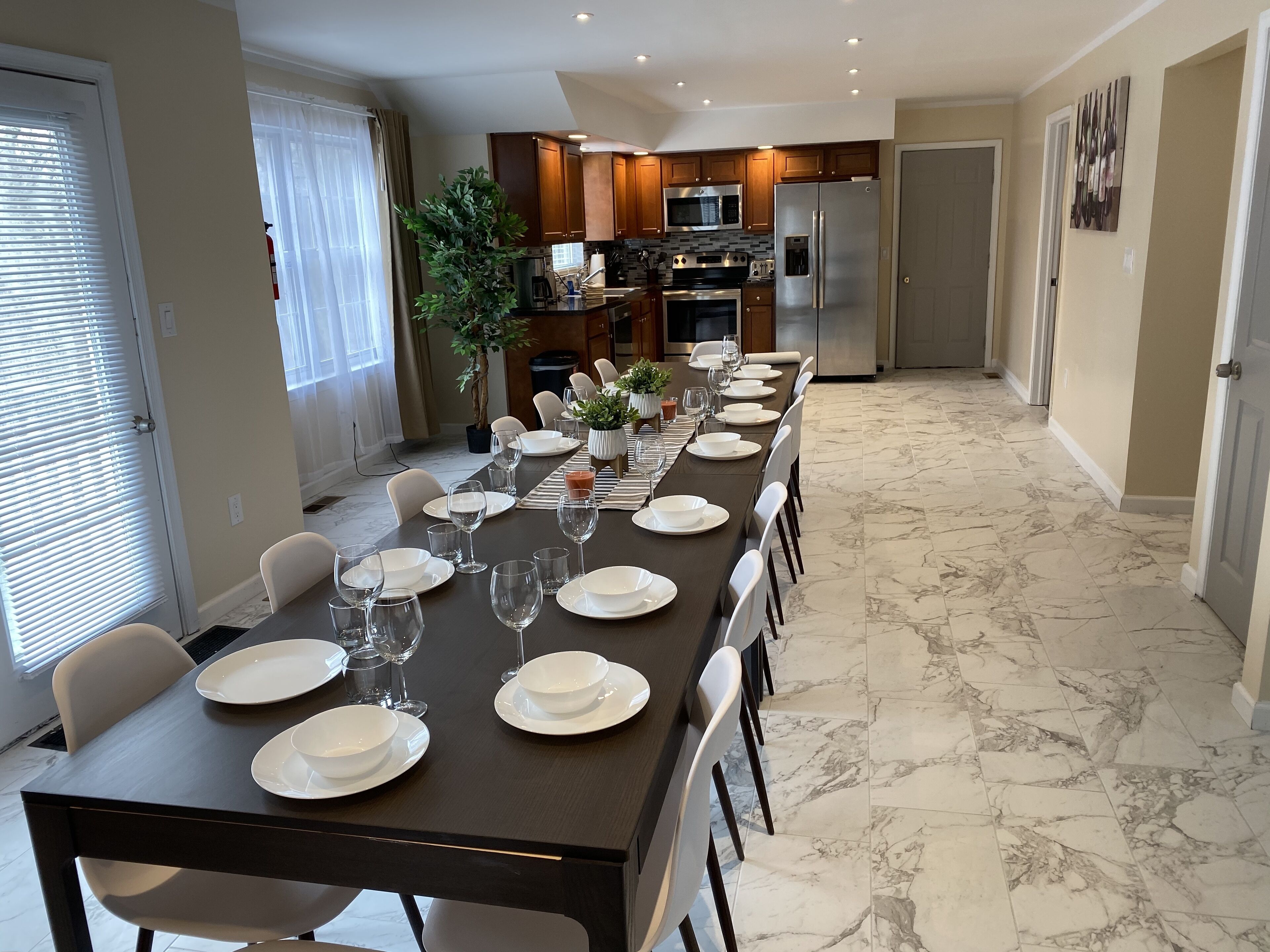 Un comedor con una larga mesa dispuesta para seis personas, rodeada de sillas, en una cocina con armarios de madera y un refrigerador de acero inoxidable.