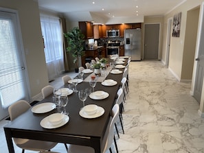 Un comedor con una larga mesa dispuesta para seis personas, rodeada de sillas, en una cocina con armarios de madera y un refrigerador de acero inoxidable.