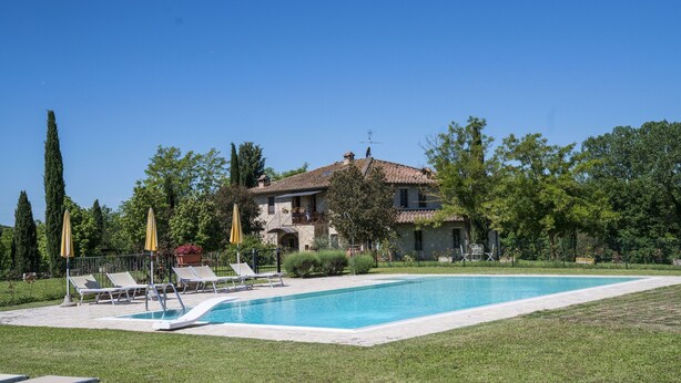 Un área junto a la piscina con sillones de descanso y sombrillas, un edificio de piedra con techo de tejas y un cielo azul despejado.