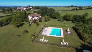 Una casa grande con piscina rodeada de sillas de jardín y sombrillas, ubicada en zona rural con colinas ondulantes y vegetación escasa.