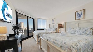 Quarto de hotel com uma cama grande, uma cama menor, uma escrivaninha com luminária, um ventilador e vista para o oceano.