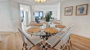 Uma sala de jantar com mesa de vidro cercada por cadeiras de vime, luminária pendente e quadros com estampas botânicas na parede.