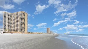 Um hotel à beira-mar com estrutura imponente, praia de areia e um cais ao longe.