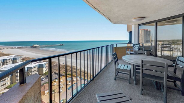 Varanda com vista para a praia e oceano, mesa com cadeiras e guarda-corpo.